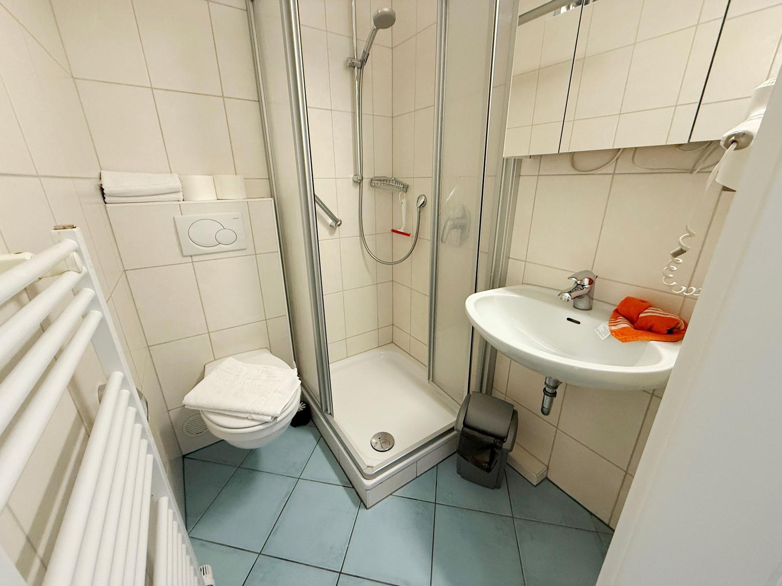 Badezimmer mit Dusche, Waschbecken und Toilette. Fliesenboden und Spiegel.