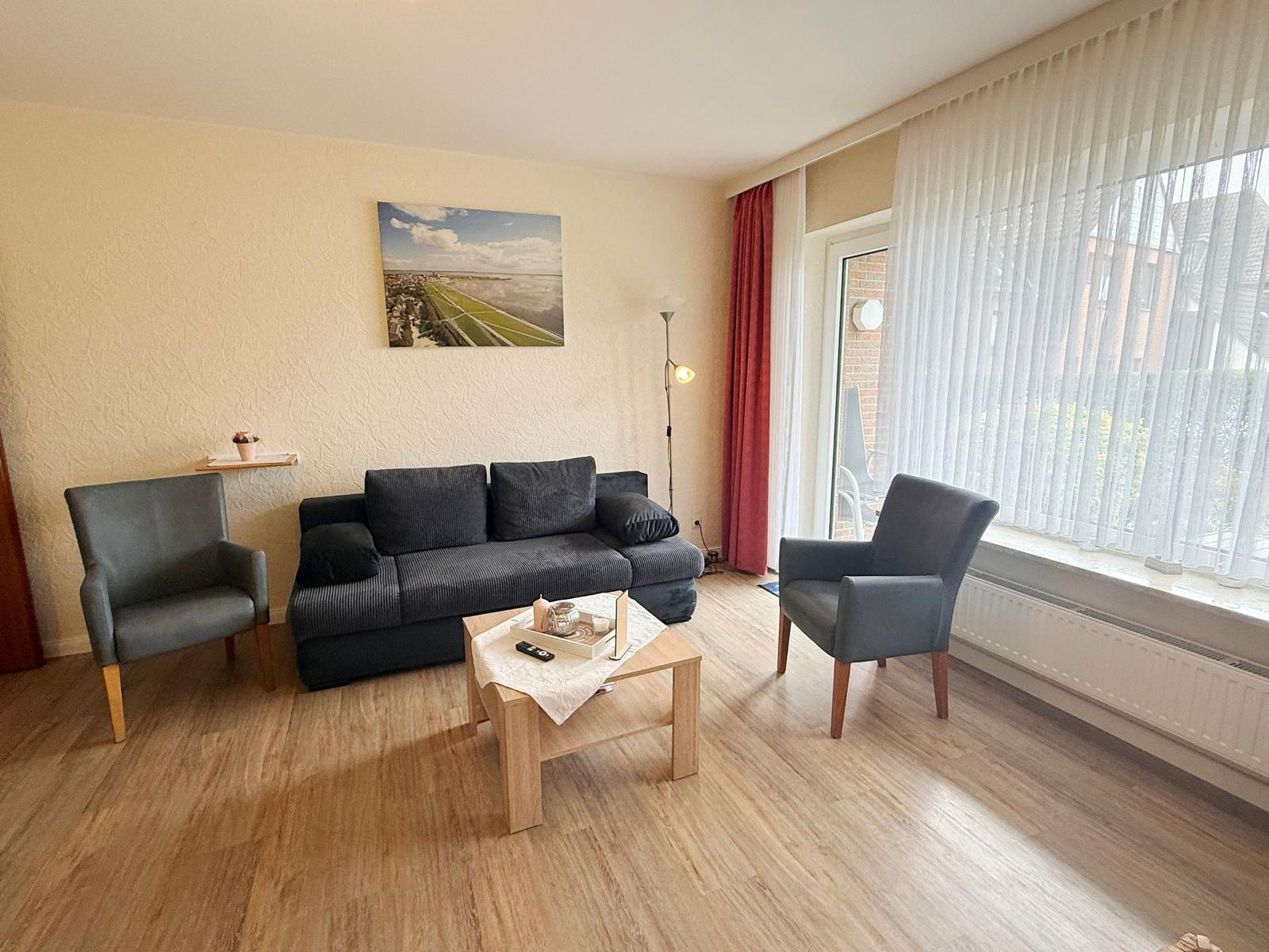 Wohnzimmer mit Sofa, zwei Sesseln, Tisch und Fenster mit Jalousien.