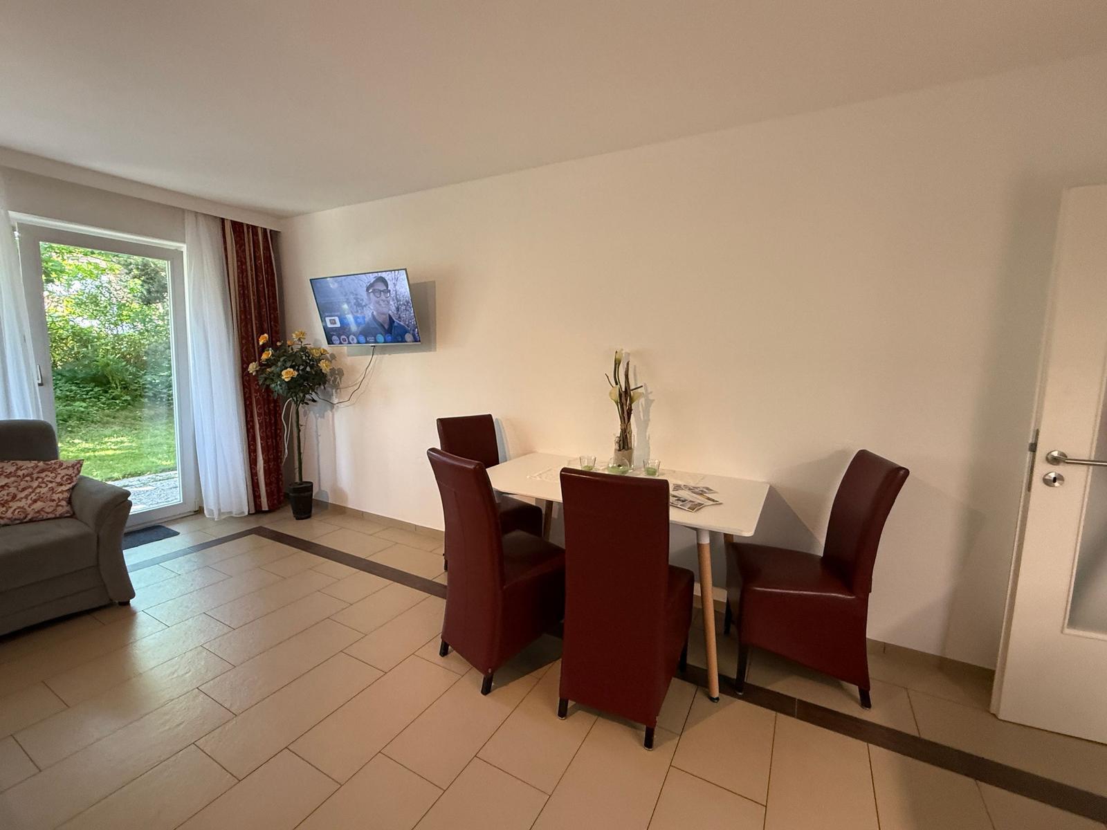 Essbereich mit Tisch und vier Stühlen, Fernseher an der Wand, Schlafzimmer im Hintergrund.