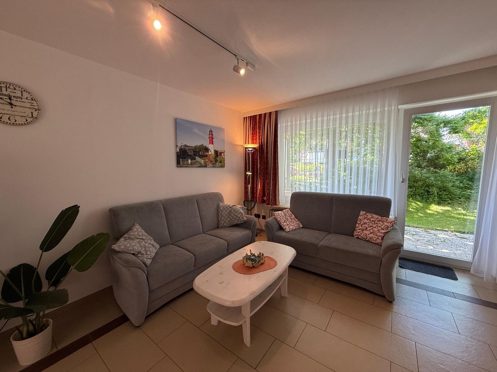 Zwei graue Sofas, ein weißer Couchtisch, eine Pflanze und ein Fenster mit Blick auf den Garten.