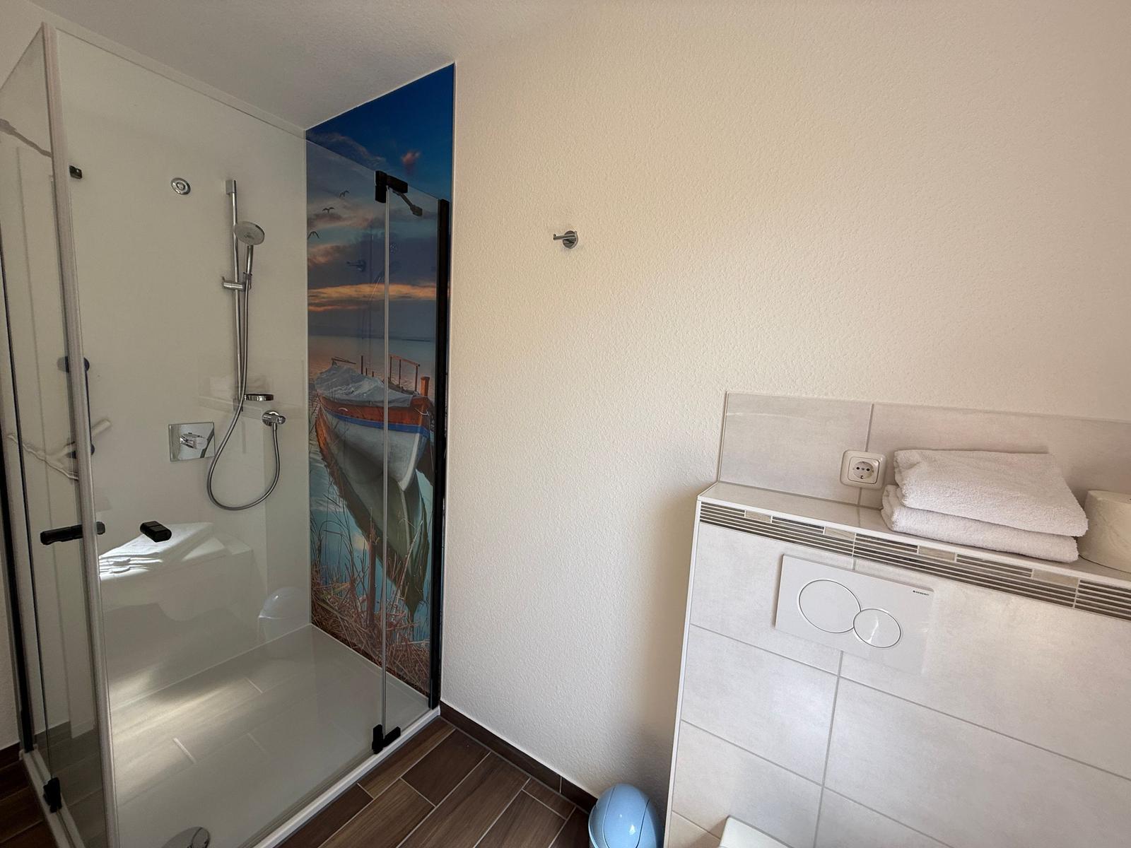 Dusche mit Glaswand und Boots-Bild. Toilette mit Handtuch und Steckdose.