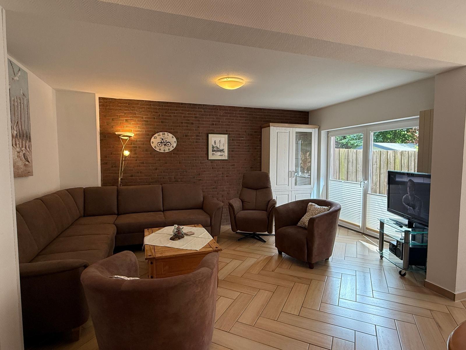 Wohnzimmer mit braunen Sitzmöbeln, Holzboden und Fernseher. An der Wand hängt eine Uhr.