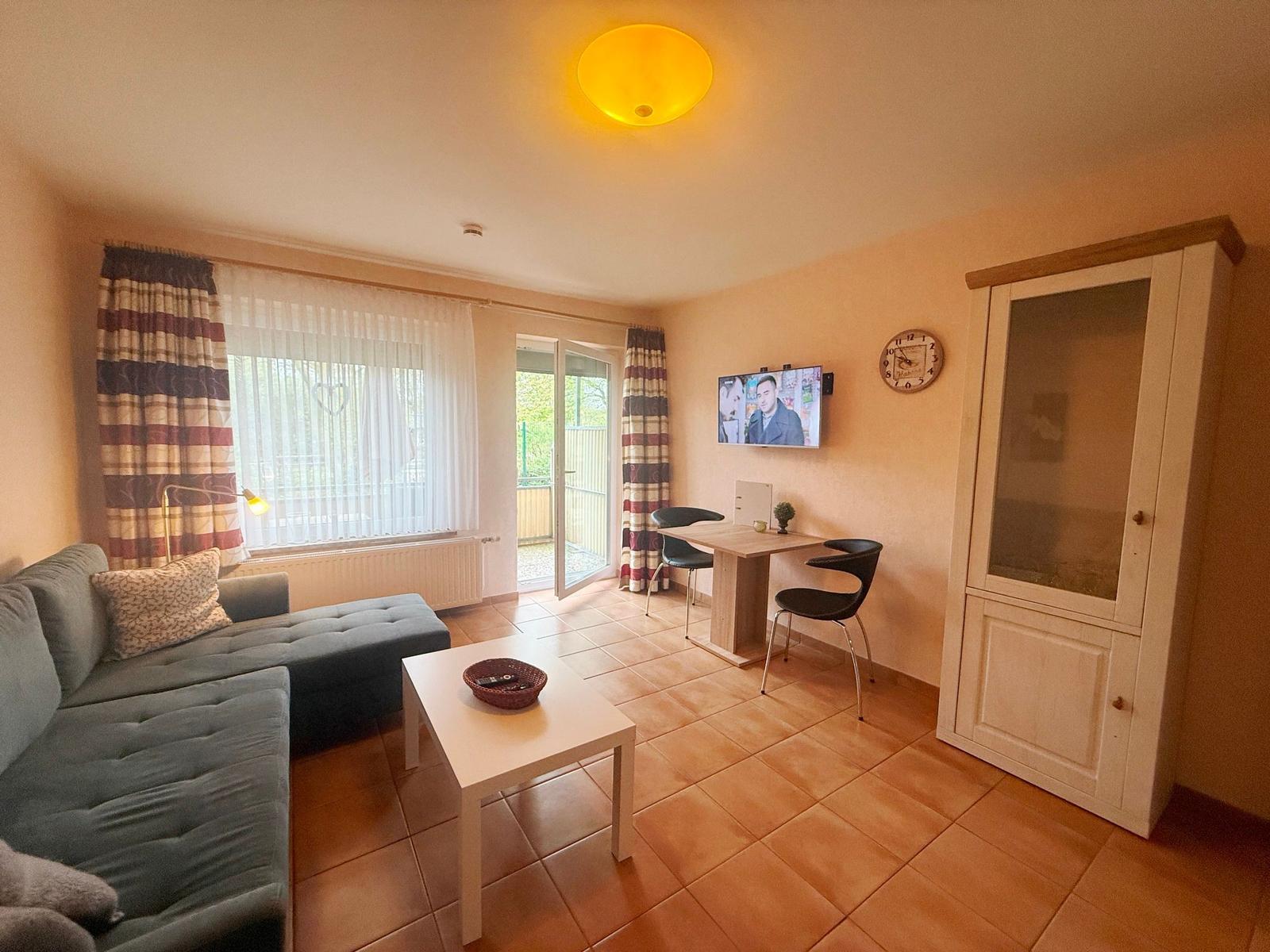 Wohnraum mit Sofa, Tisch, TV und Balkontür. Fliesenboden und Holzschrank.