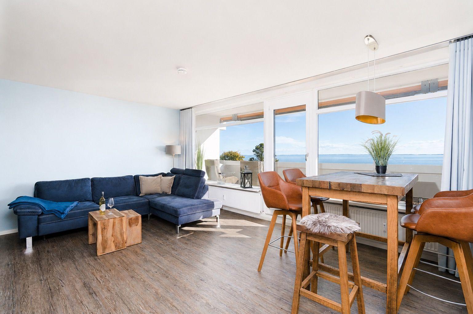 Wohn- und Essbereich mit Blick aufs Meer. Blauer Sofa, Holztisch und braune Stühle.