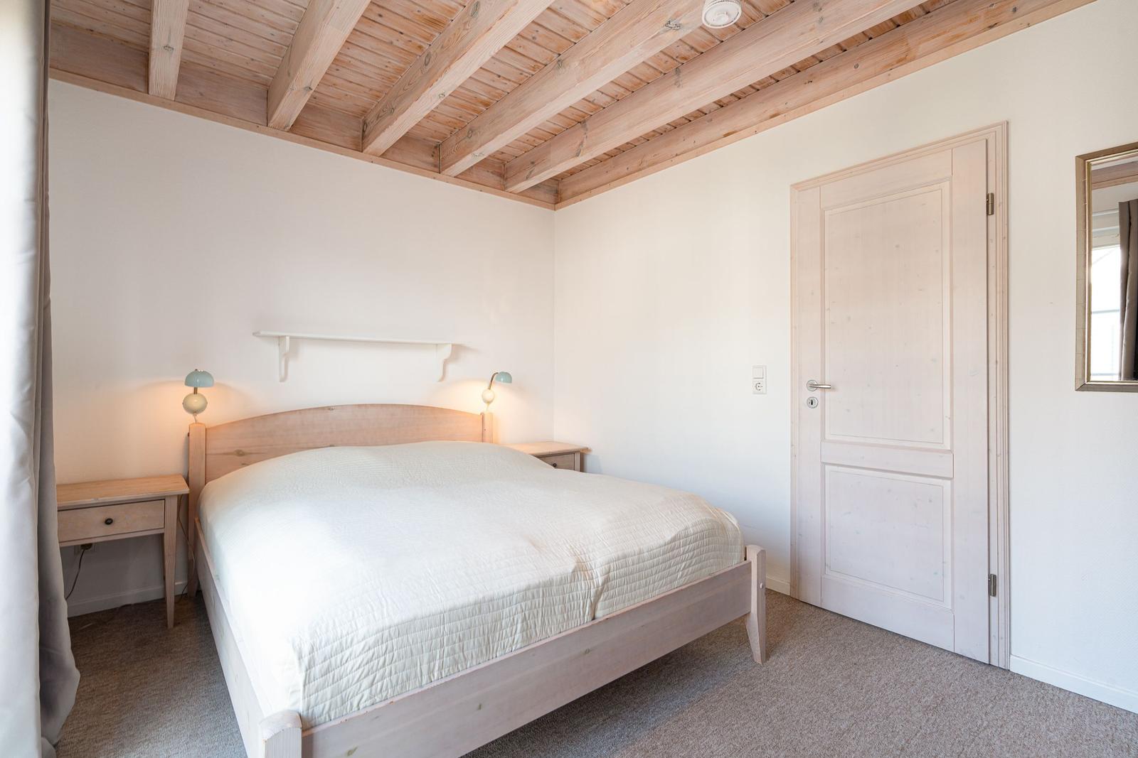 Bedroom with wooden floor, bed, and nightstands. Ceiling beams and door are visible.
