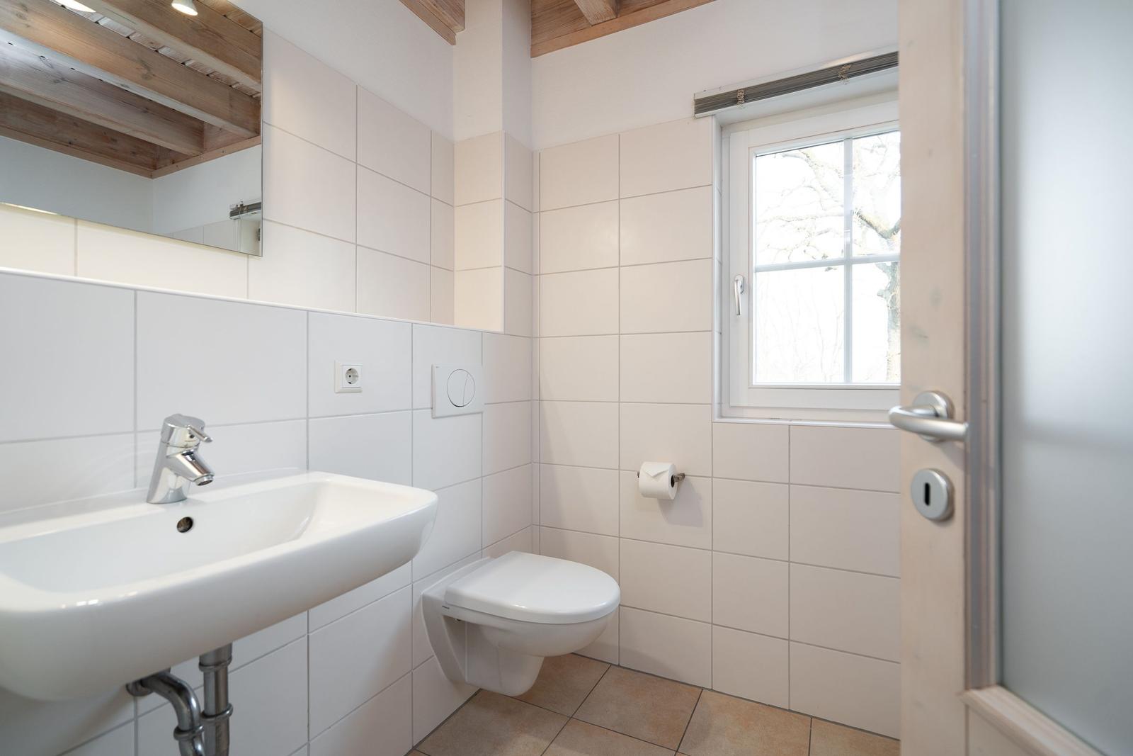 Bathroom with sink, toilet, and window. White tiles and wooden beams on ceiling.