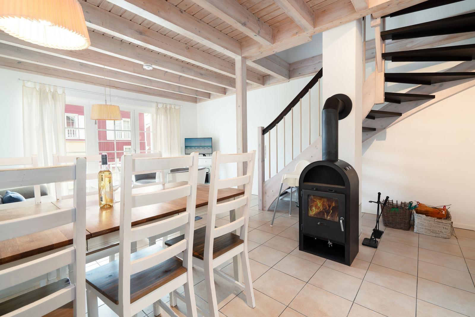 Dining room with wooden table, white chairs, and wood stove. Stairs lead upstairs.