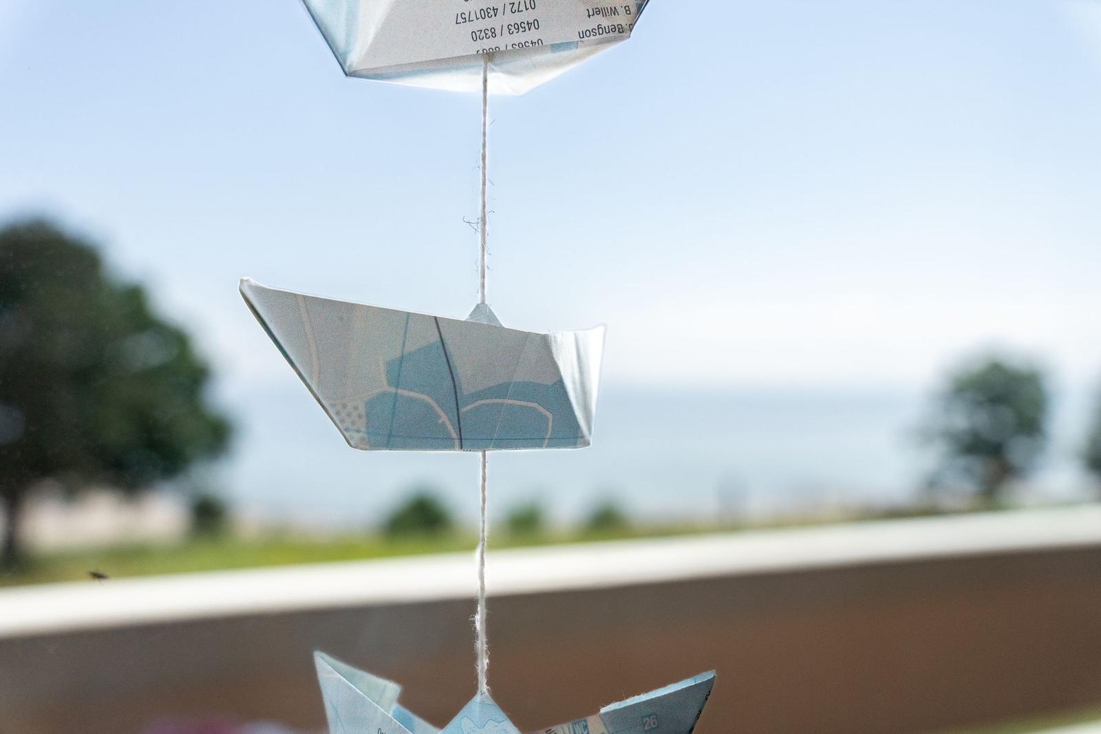 Origami-Schiff hängt an einem Faden vor einem Fenster mit Blick auf Grünflächen.