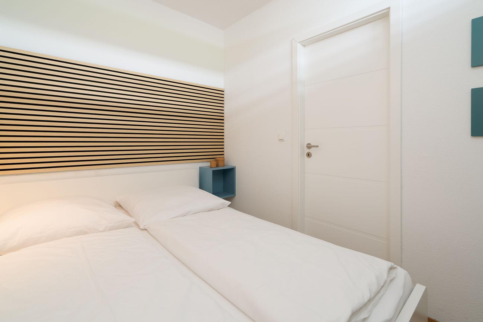 Schlafzimmer mit Bett, Kopfteil aus Holzstreifen und blauer Nachttischkante.