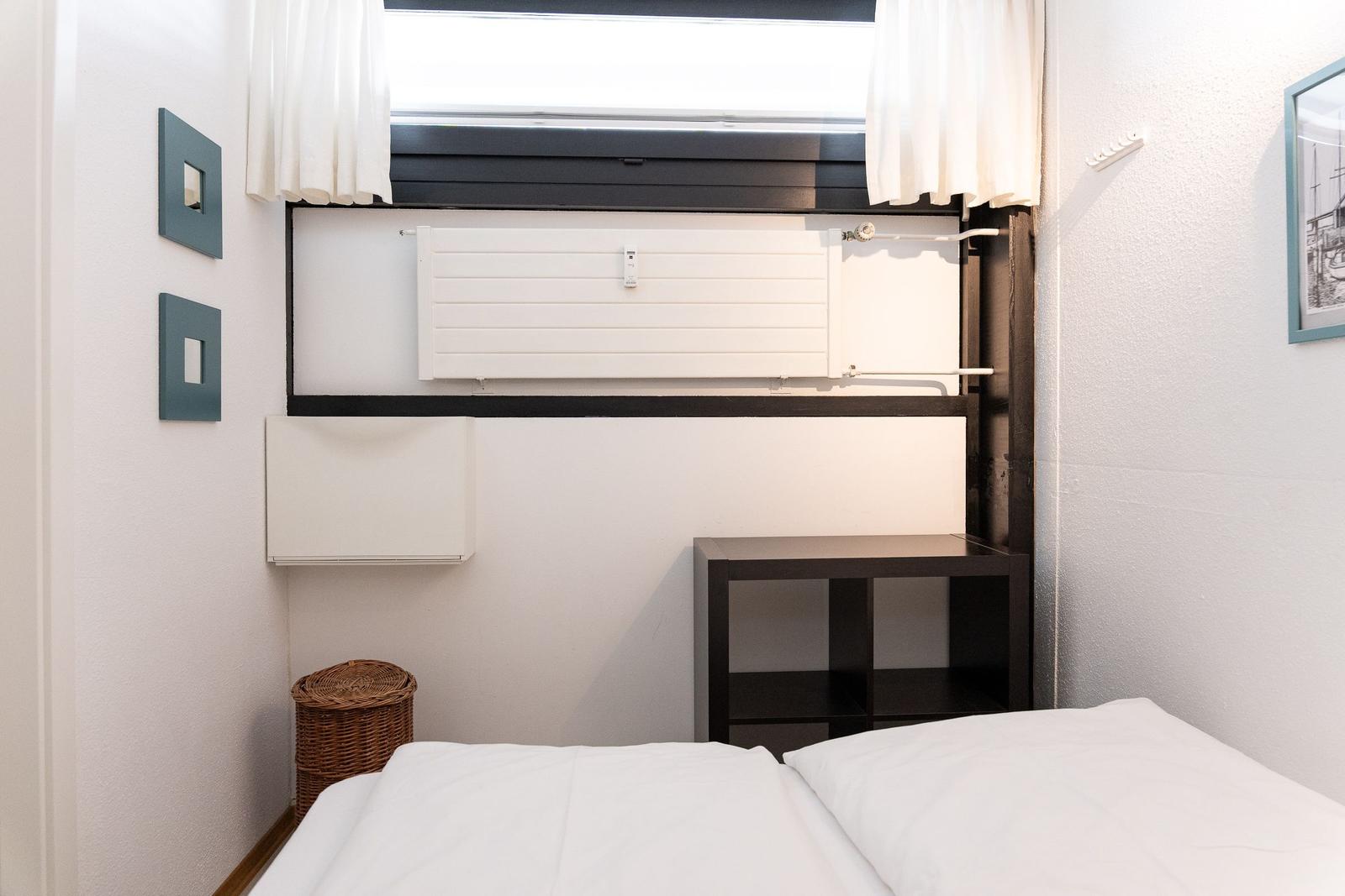 Ein Bett mit weißer Bettwäsche, ein dunkler Schrank und ein Fenster mit weißen Vorhängen.