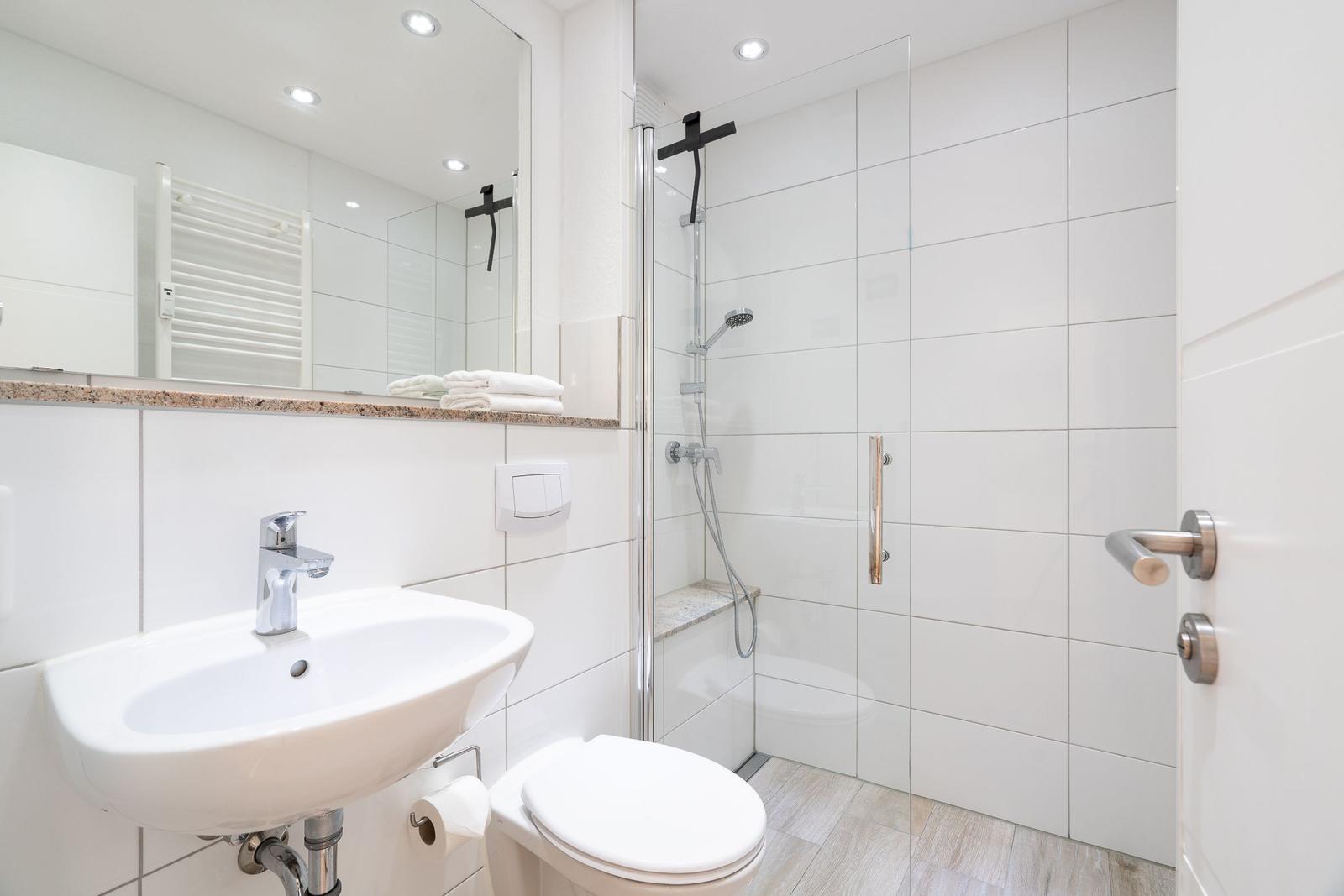 Badezimmer mit Dusche, Waschbecken und Toilette. Weiße Fliesen und Spiegel.