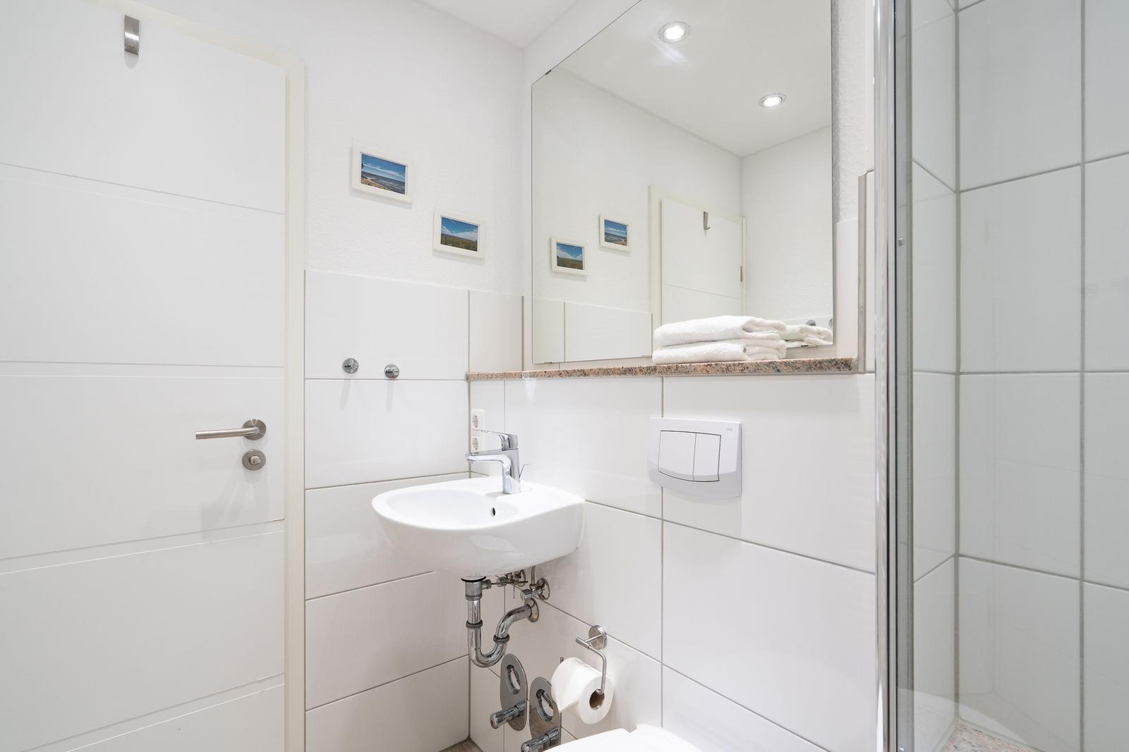 Badezimmer mit Waschbecken, Spiegel und Dusche. Weiße Fliesen und Handtuch auf Ablage.