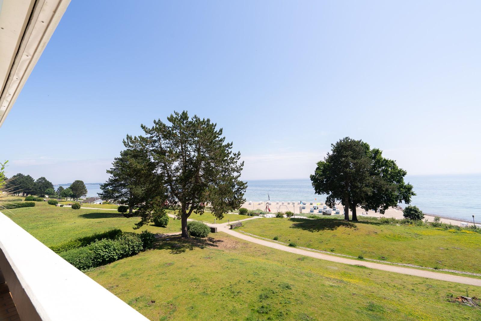 Blick vom Balkon auf Grünfläche, Bäume und Strand mit Meer im Hintergrund.
