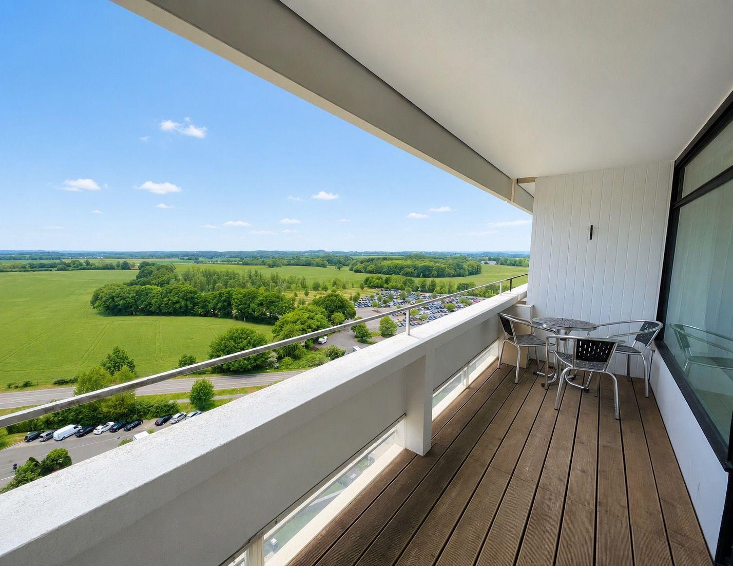 Balcony with table and chairs, view of green landscape and parking lot.