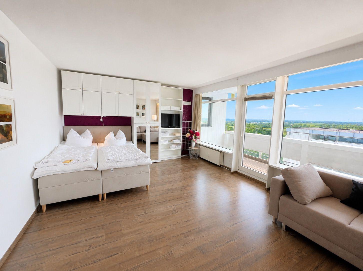 Room with double bed, sofa, and large window with view of the landscape.