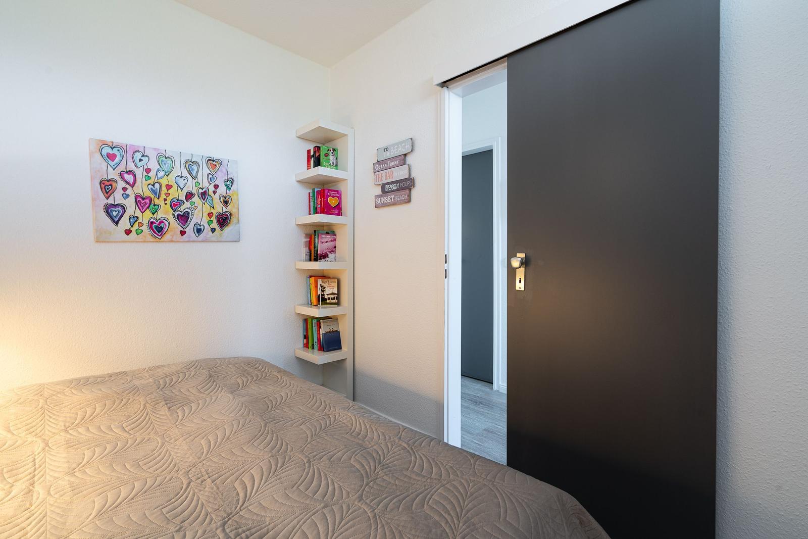 Schlafzimmer mit Bett, Wandbild und Bücherregal. Tür führt in einen Flur.