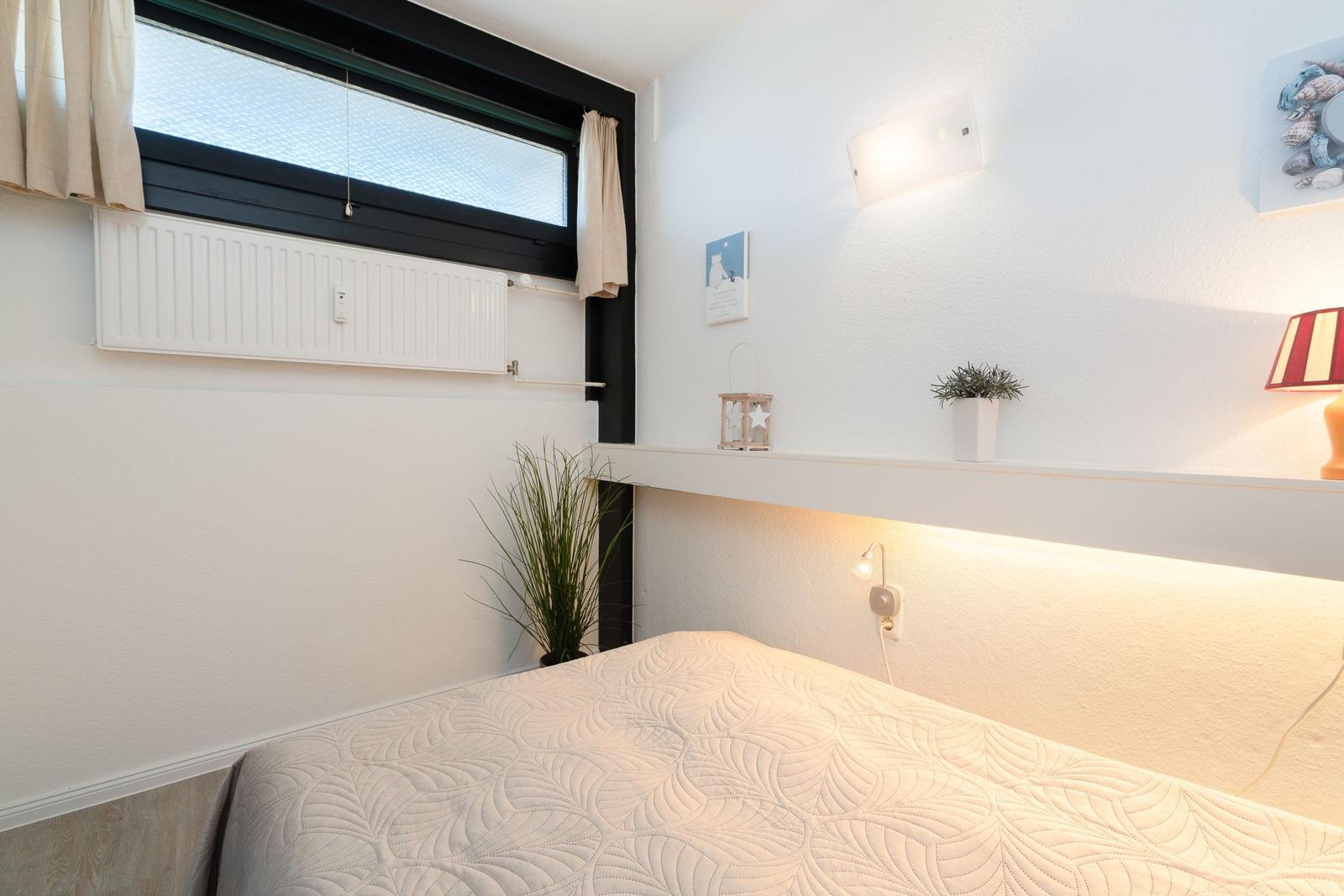 Schlafzimmer mit Bett, Fenster und Wandregal mit Pflanzen und Lampen.