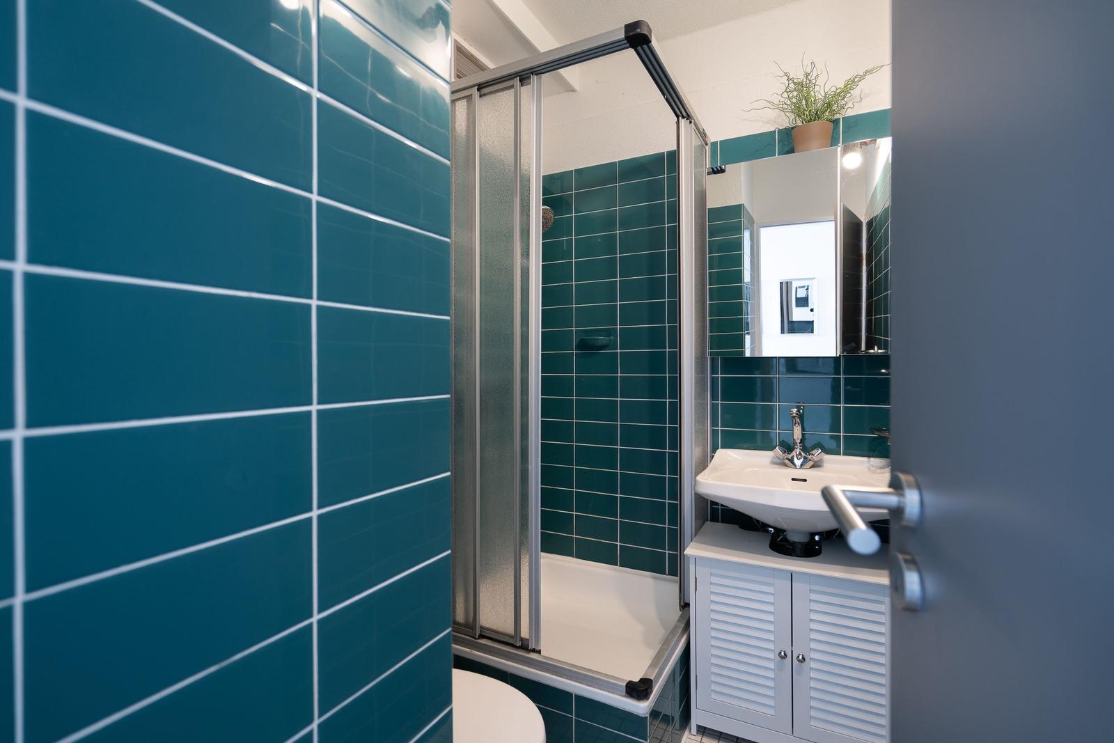Badezimmer mit Dusche, Waschbecken und Toilette. Wände mit türkisen Fliesen verkleidet.