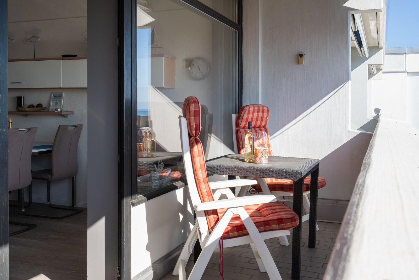 Terrasse mit Tisch, zwei Stühlen und Blick aufs Meer. Im Hintergrund Küche und Wanduhr.