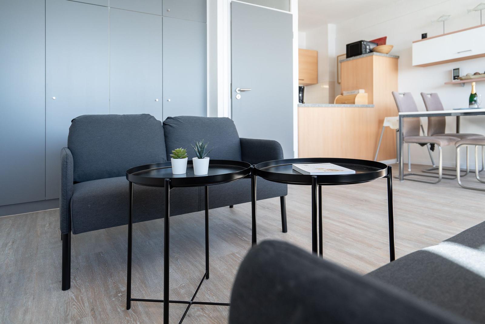 Ein Wohnbereich mit grauem Sofa, zwei schwarzen Tischen und Pflanzen. Im Hintergrund Küche und Essbereich.