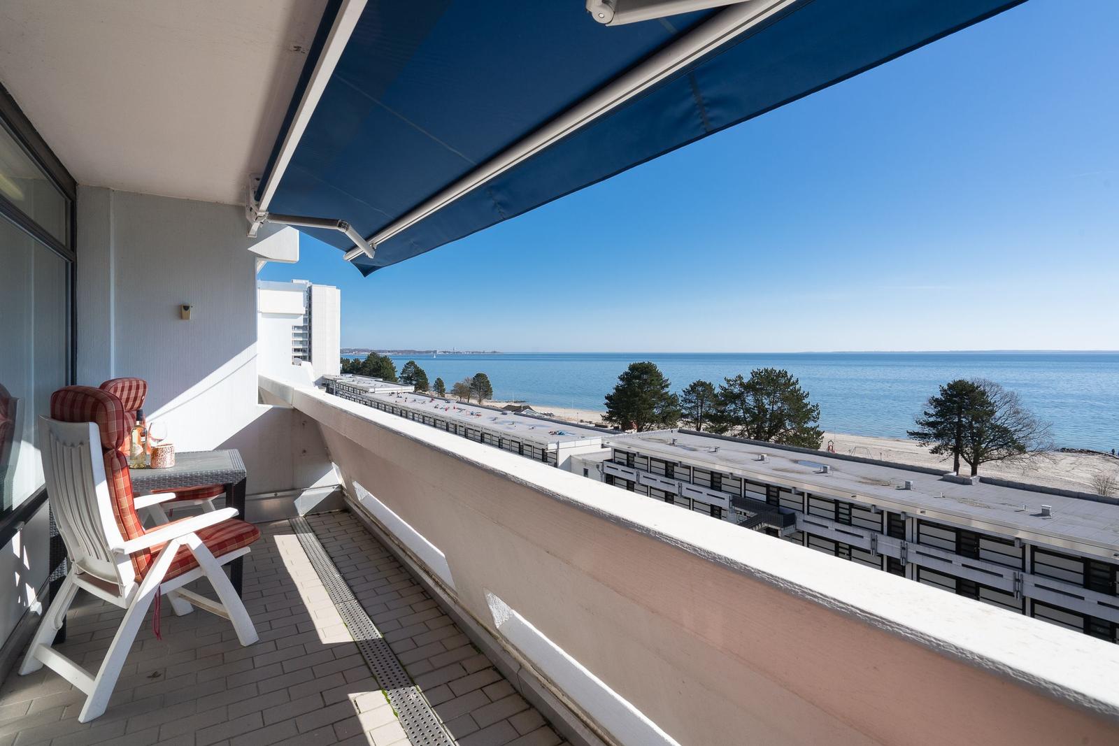 Terrasse mit Blick auf Meer und Strand. Blauer Sonnenschutz und weiße Gartenstühle.