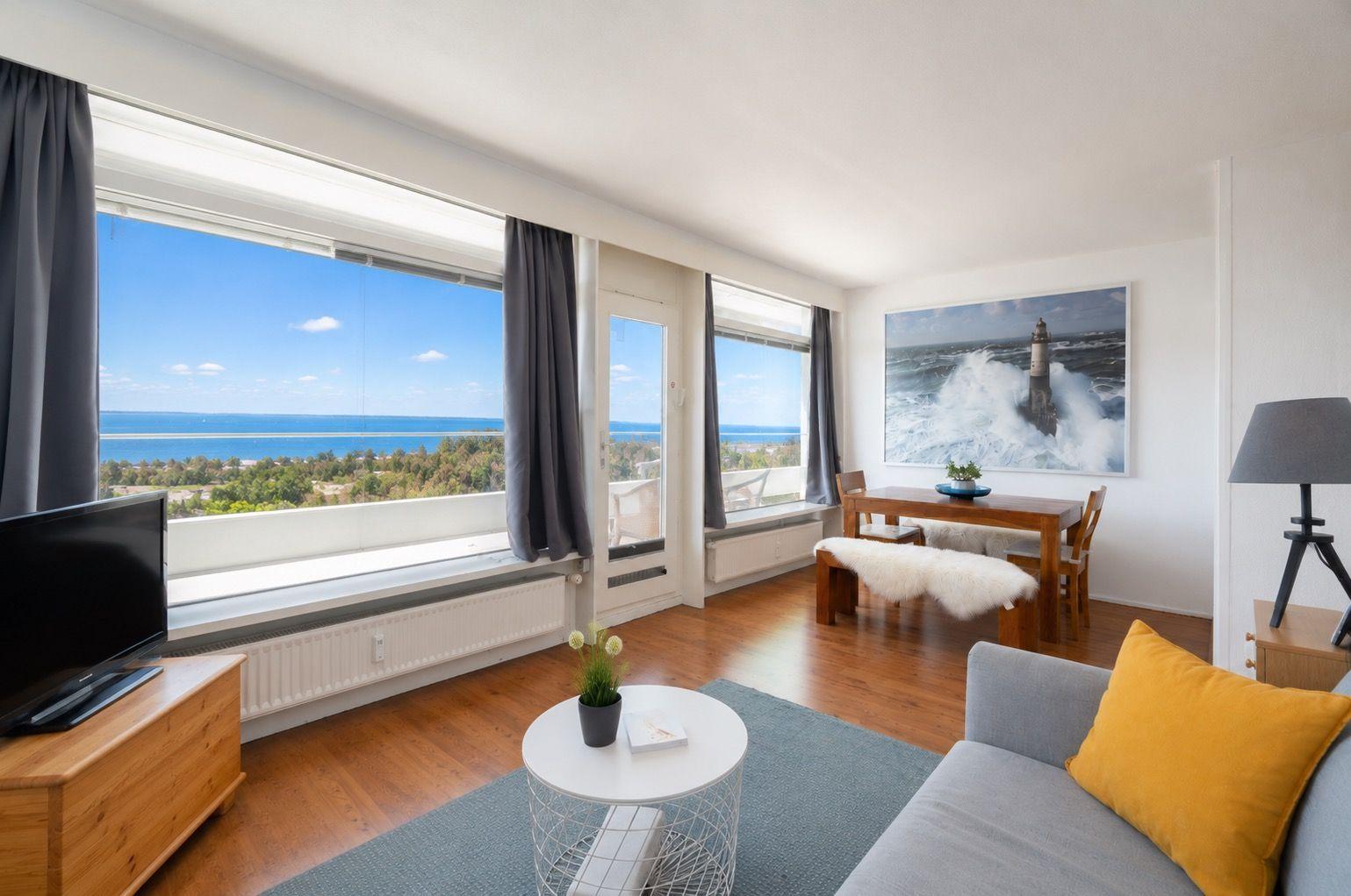 Apartment with sea view, dining table, and sofa. Large windows and wall art with lighthouse.