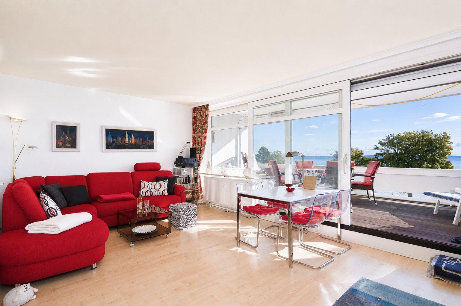 Living and dining area with red sofa, glass table, and balcony view of the sea.