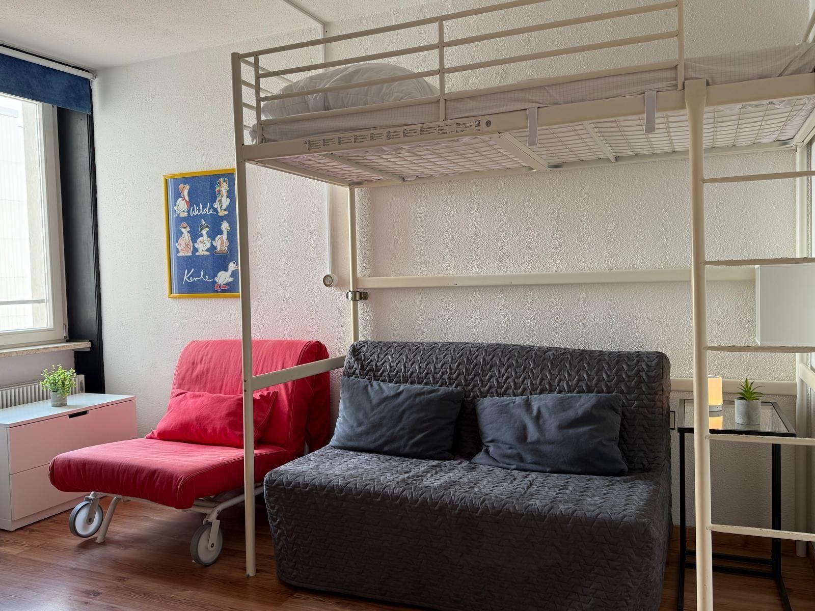 Ein Raum mit einem Hochbett, einem grauen Sofa und einem roten Klappstuhl.