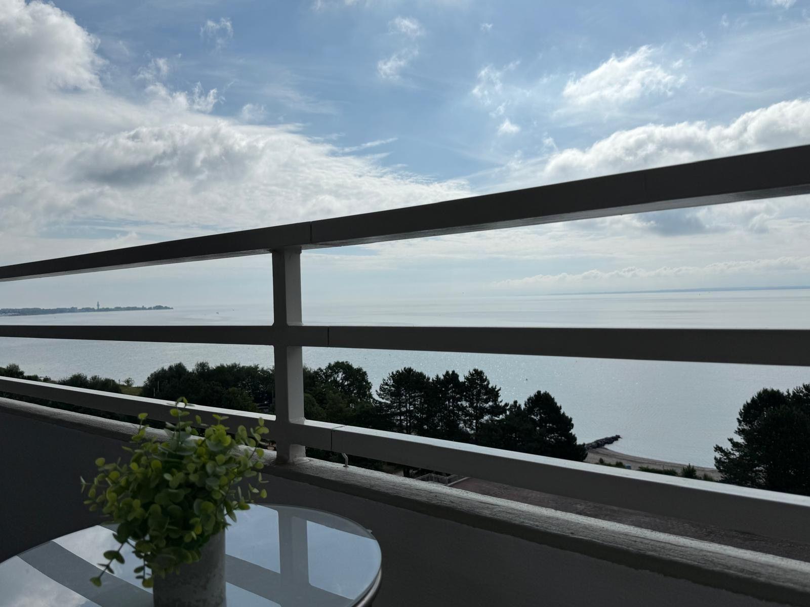 Balcony with sea view, small table and plant. Trees and horizon visible.