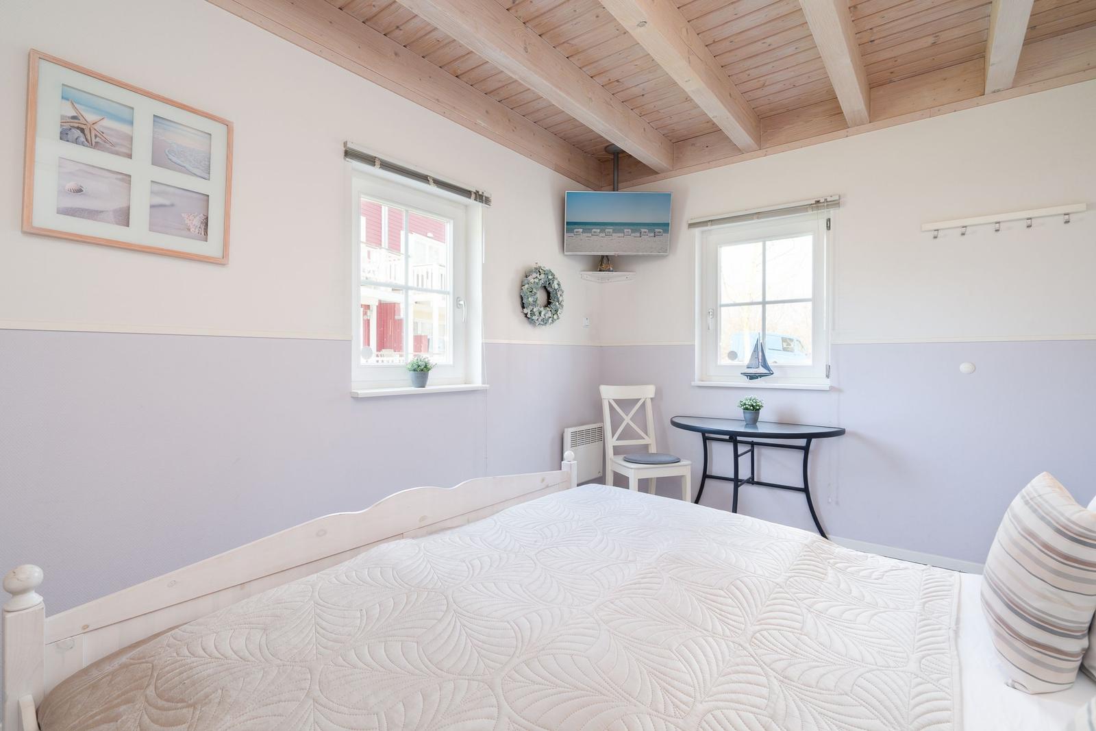 Schlafzimmer mit Bett, Tisch, Stuhl und Fenstern. Holzdecke und Wandgestaltung.