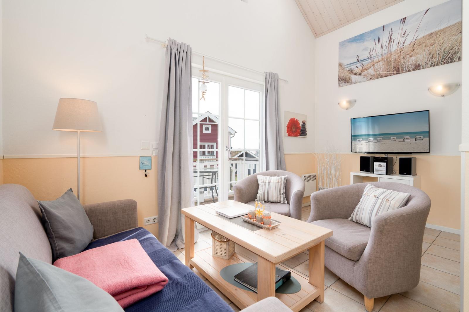 Wohnzimmer mit Sofa, zwei Sesseln, Tisch und TV. Fenster mit Blick auf Balkon.