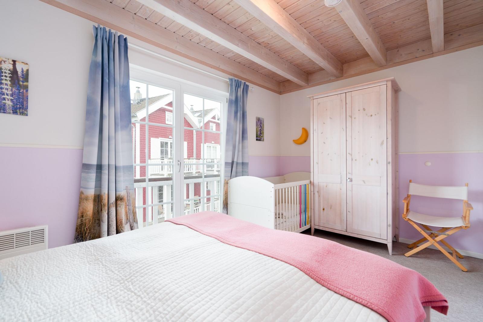 Schlafzimmer mit Bett, Kinderbett, Kleiderschrank und Fensterblick auf rotes Haus.
