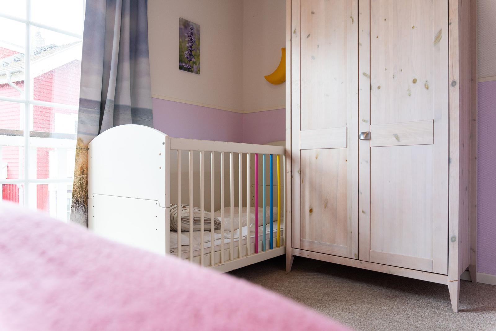 Kinderzimmer mit weißem Kinderbett und Holzschrank. Wandfarbe lila. Fenster mit Vorhang.
