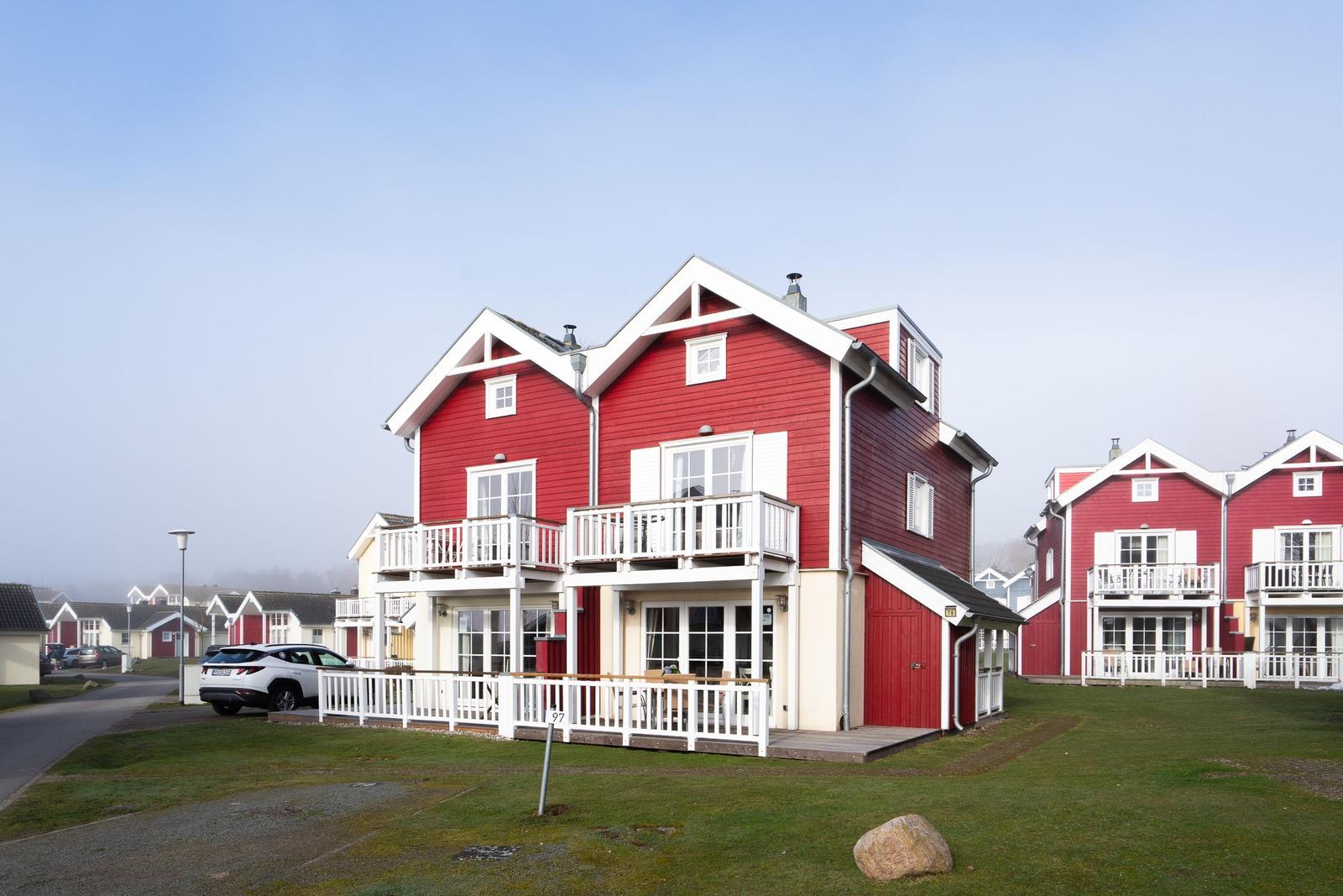 Rotes Ferienhaus mit weißen Balkonen und Terrasse. Grünflächen und Nebengebäude im Hintergrund.