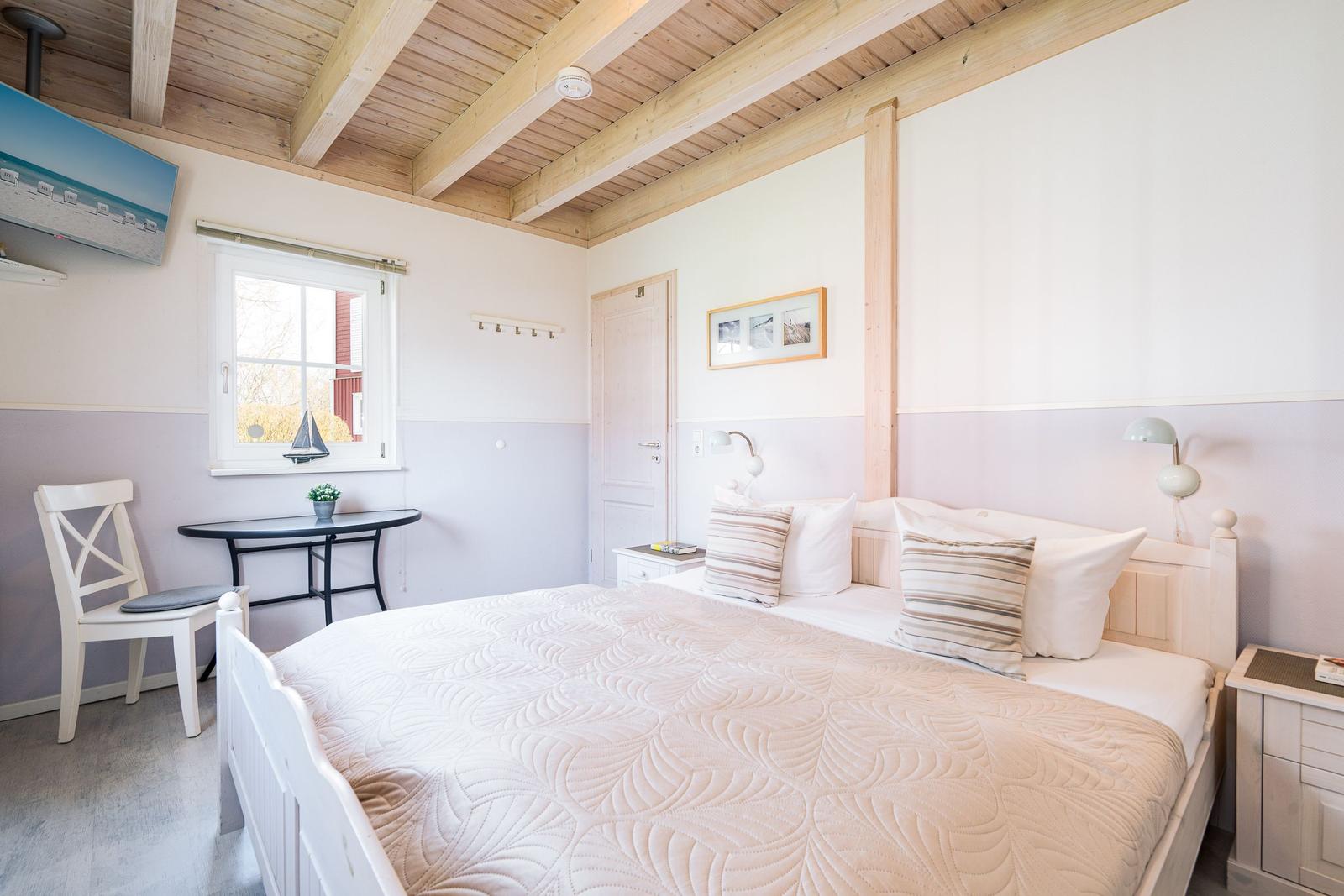 Schlafzimmer mit weißem Bett, Holzbalkendecke und kleinem Tisch mit Stuhl.
