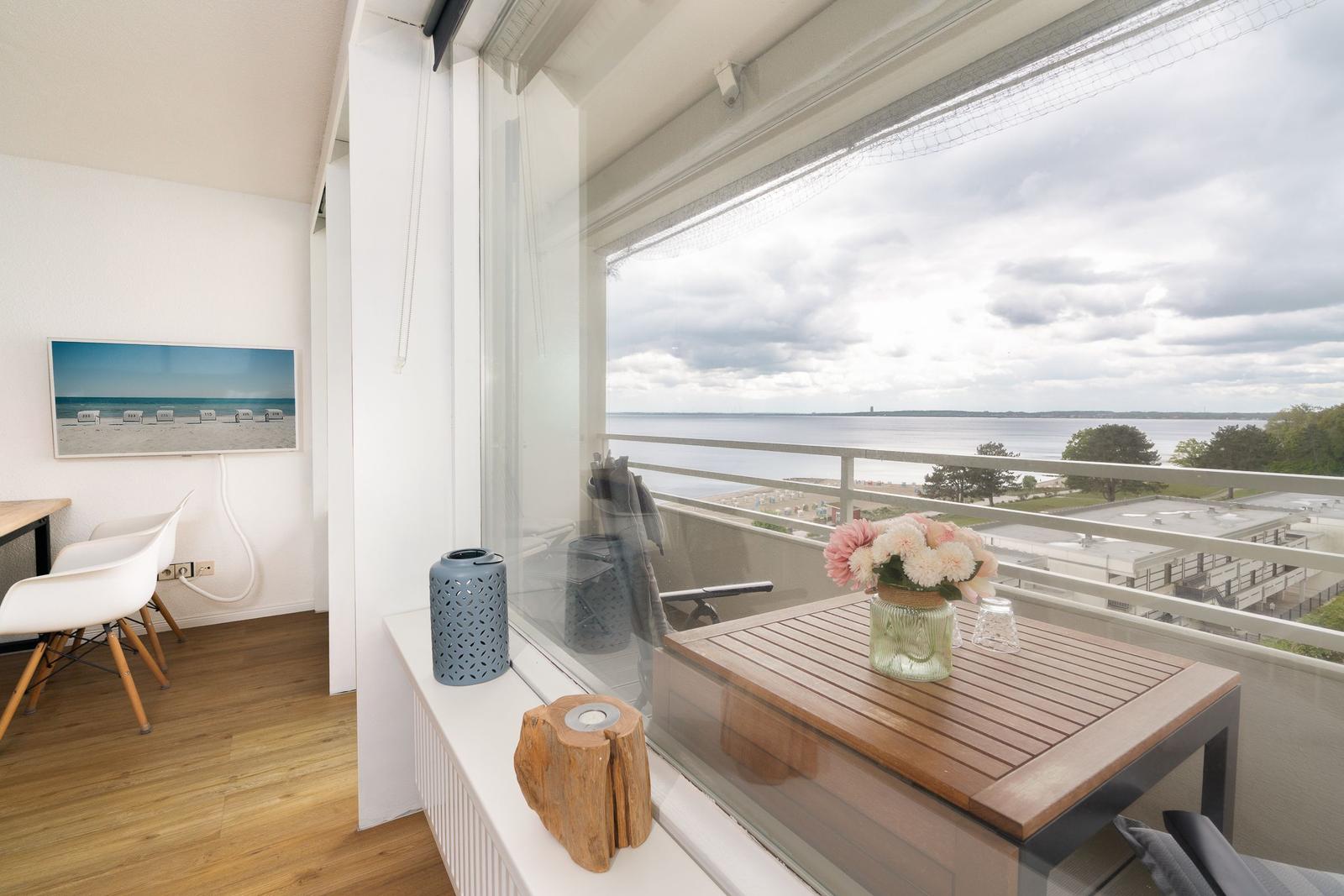 Balcony with wooden table, flowers, and sea view. Interior shows white chairs and wall art.