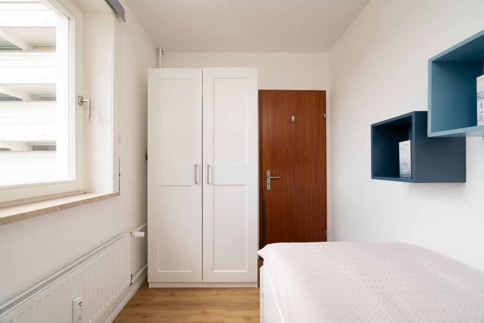 Schlafzimmer mit Bett, Kleiderschrank und Fenster. Wandregale sind blau.