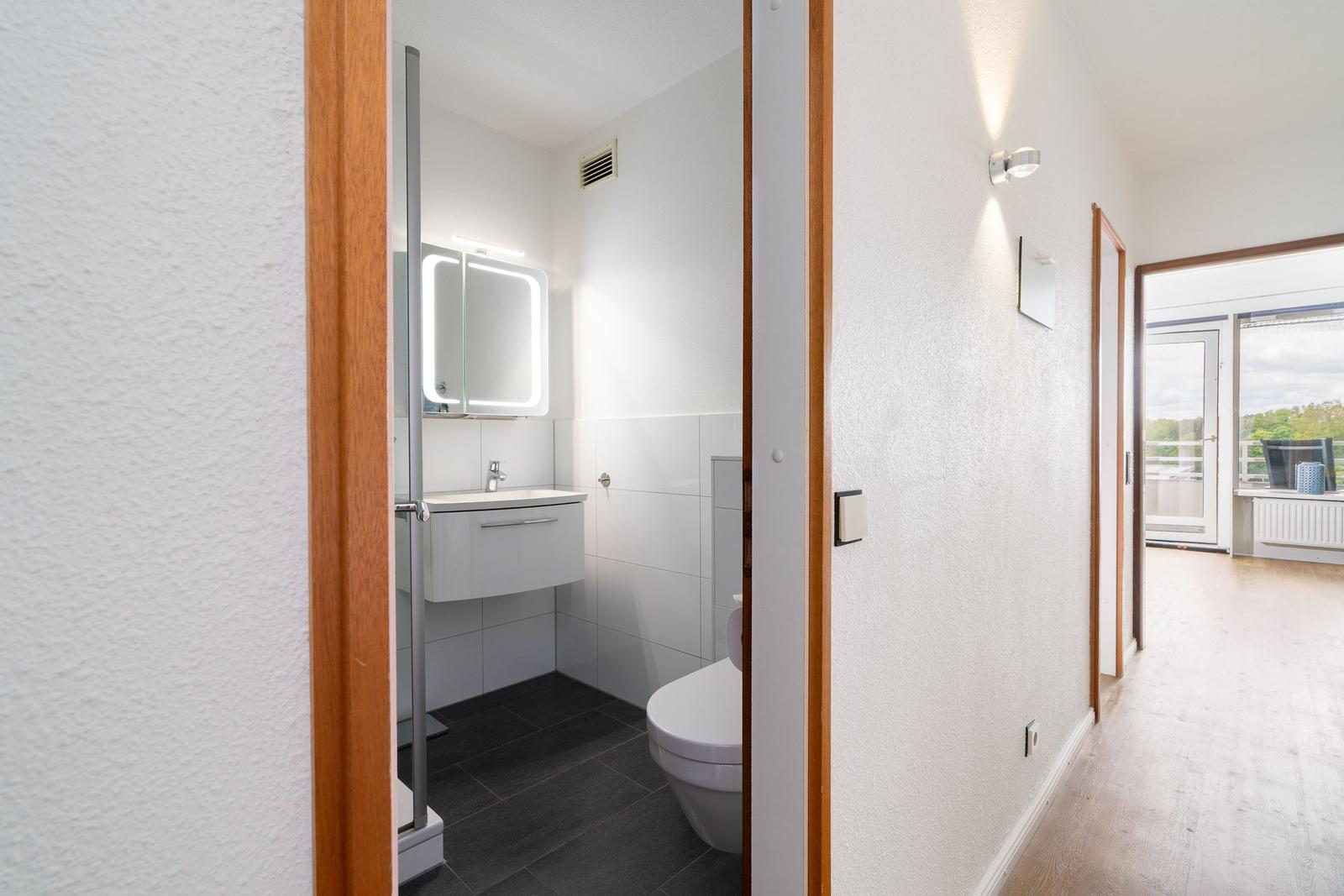 Badezimmer mit Spiegel, Waschbecken und Toilette. Flur führt zu Balkon.