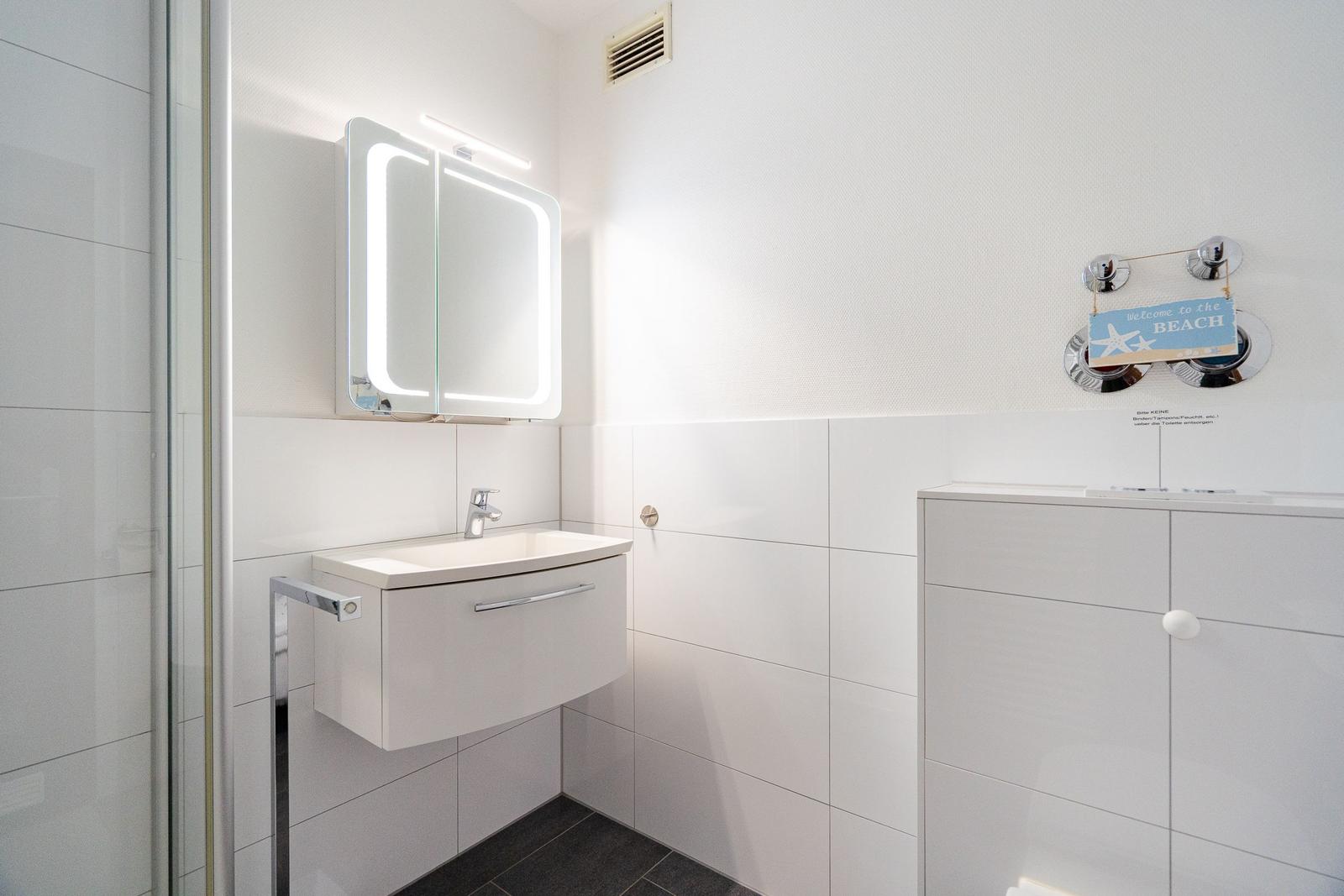 Badezimmer mit Spiegel, Waschbecken und Dusche. An der Wand hängt ein Schild mit Strandmotiv.