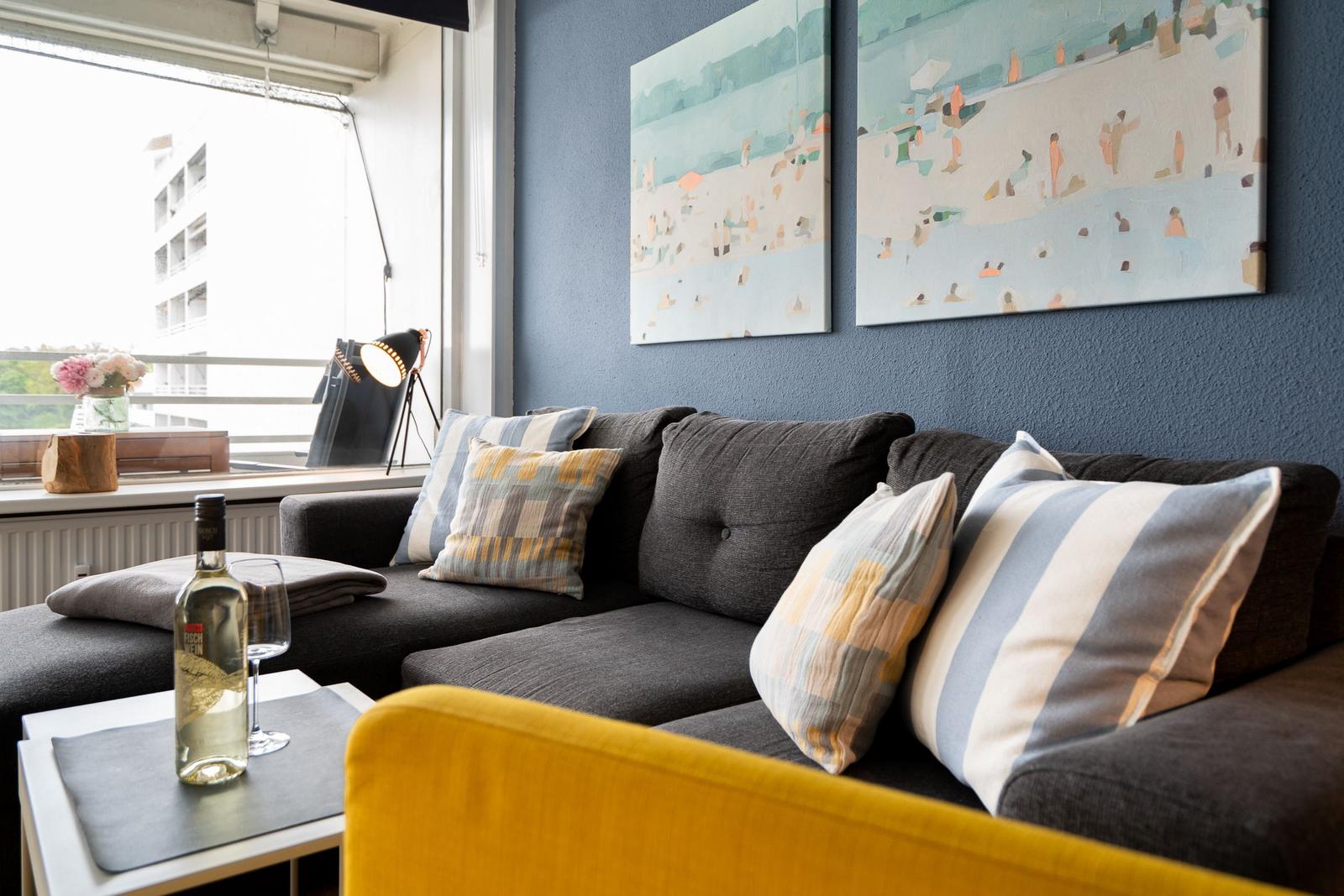 Ein Wohnzimmer mit grauem Sofa, Kissen und Wandbildern. Auf dem Tisch steht eine Weinflasche.
