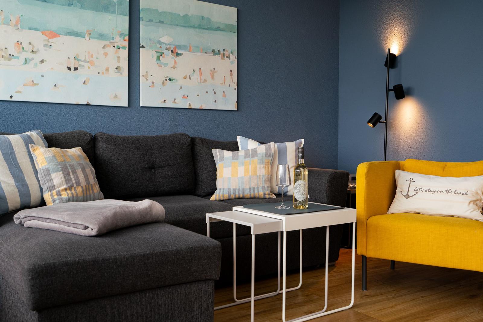 Ein Wohnbereich mit grauem Sofa, gelbem Sessel und Wandbildern an blauer Wand.