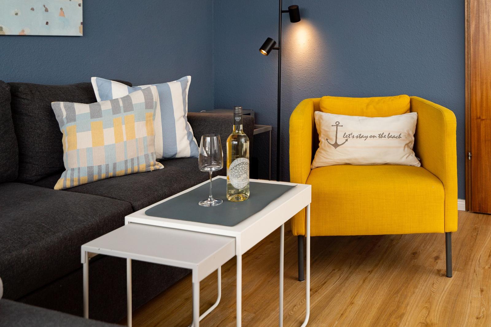 Ein Wohnbereich mit grauem Sofa, gelber Sessel und Tisch mit Weinflasche.