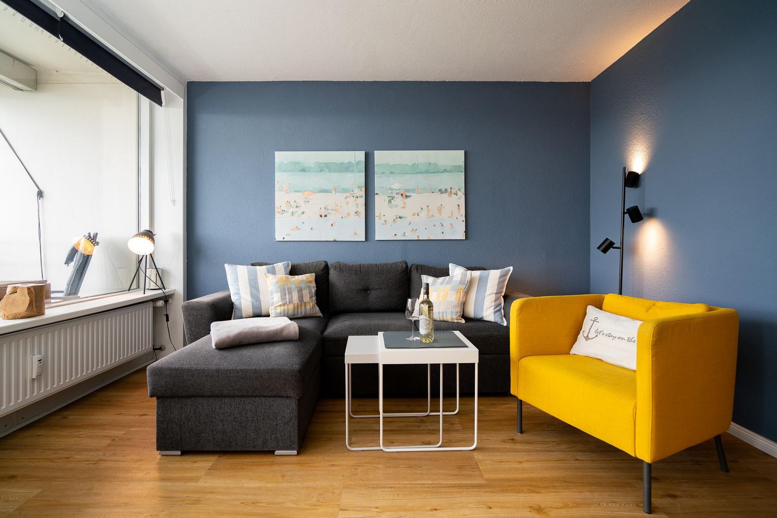 Ein Wohnzimmer mit grauem Sofa, gelbem Sessel und zwei Wandbildern an blauer Wand.