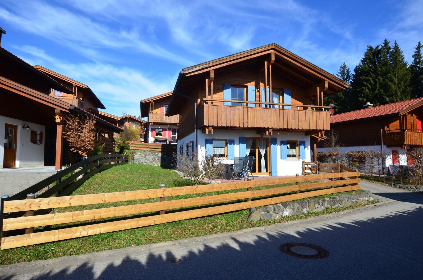 Wooden house with balcony, blue shutters, and garden fence. Backyard with terrace and chairs.