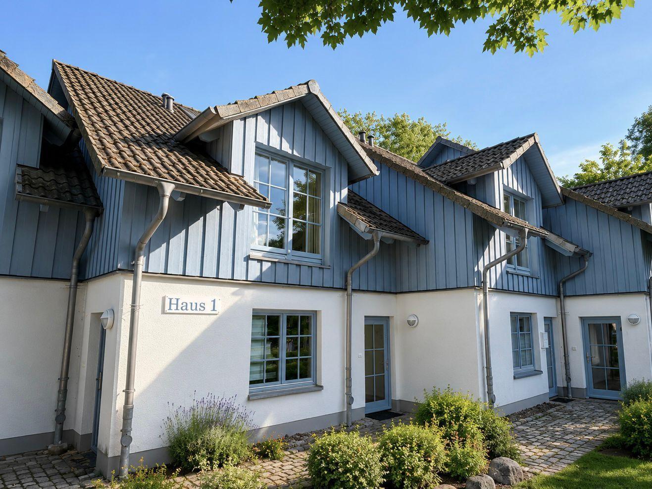 Haus 1 mit blauen Holzfassade und weißen Wänden. Mehrere Fenster und Türen. Grünflächen und Steinweg.