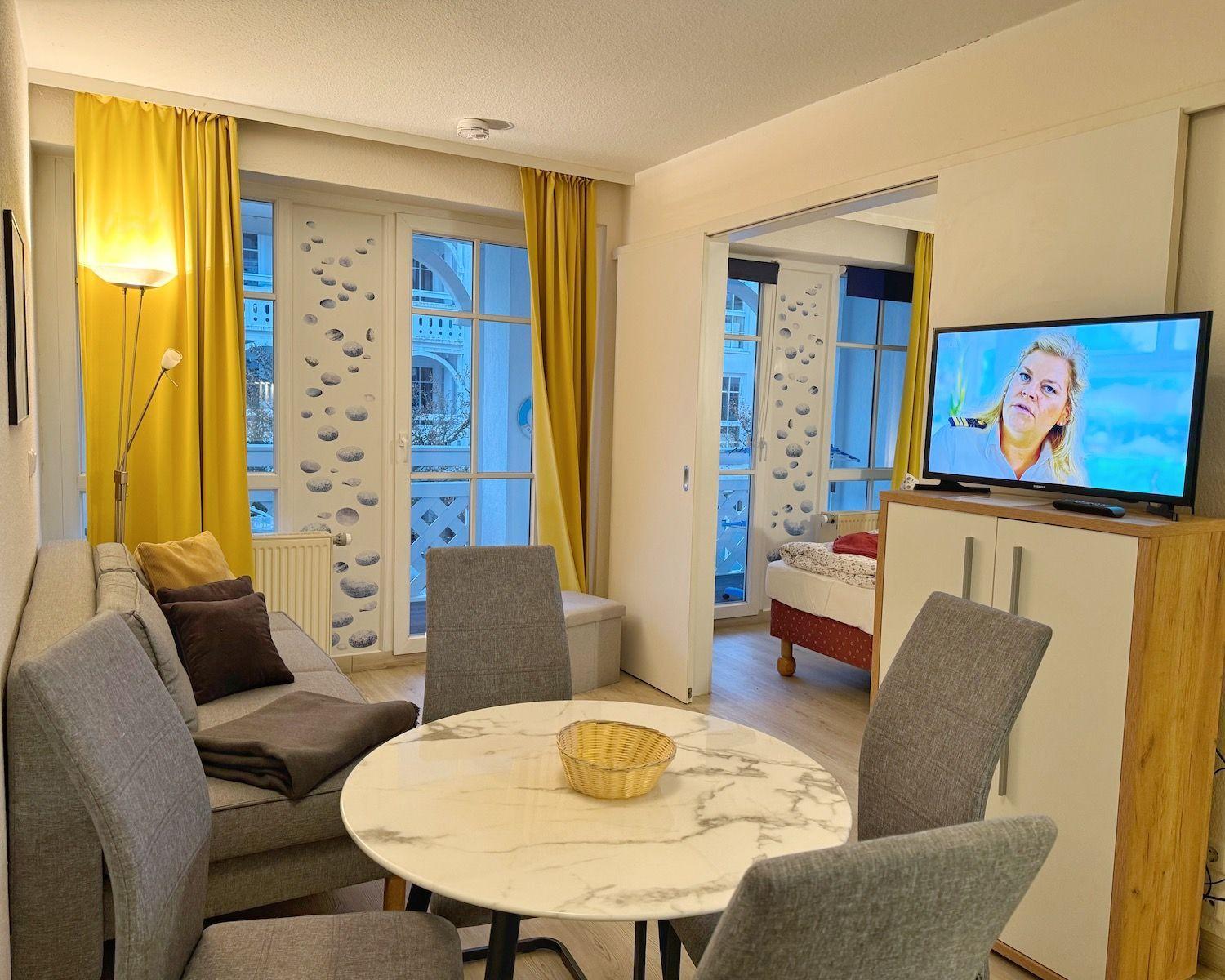 Living area with dining table, sofa, and TV. Passage to bedroom. Yellow curtains.