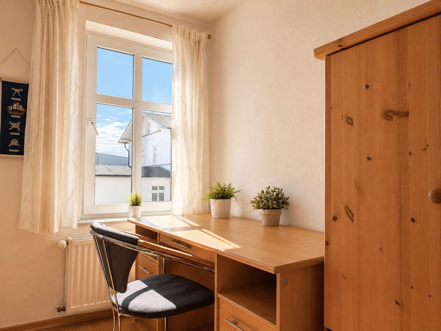 Schreibtisch mit Stuhl und Pflanzen neben Fenster und Kleiderschrank.