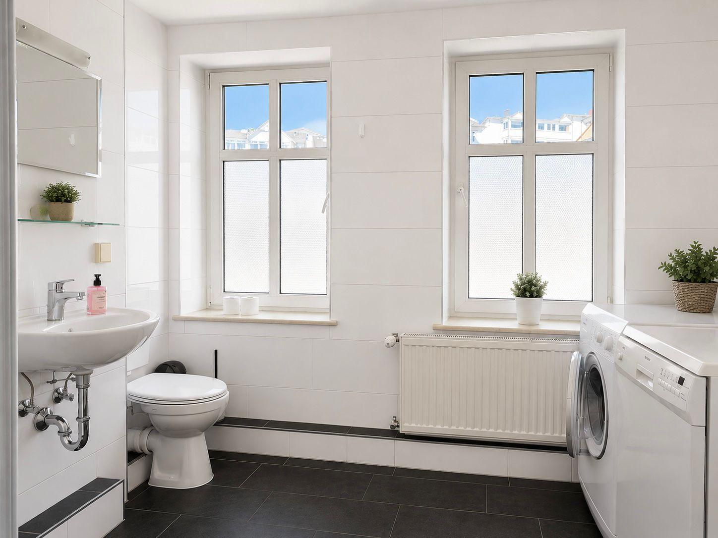Badezimmer mit Toilette, Waschbecken und Fenstern. Weiße Wände, schwarze Fliesen und Waschmaschine.