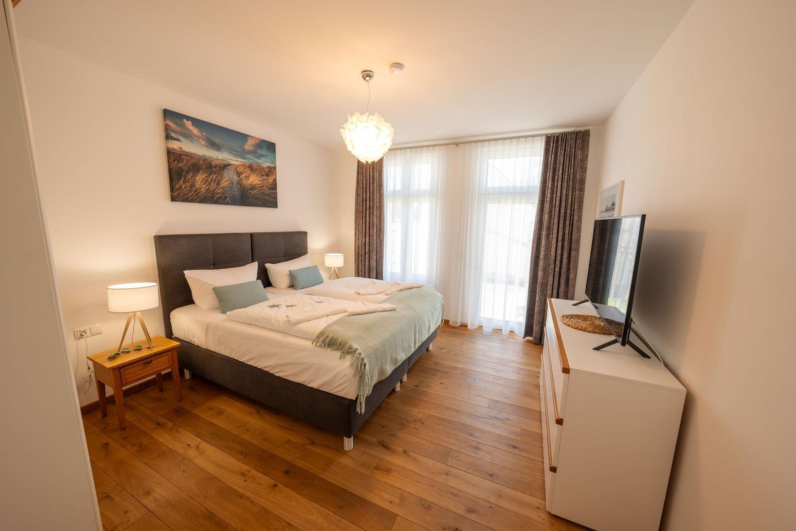 Schlafzimmer mit Doppelbett, Holzboden, TV und Fenster mit Vorhängen.