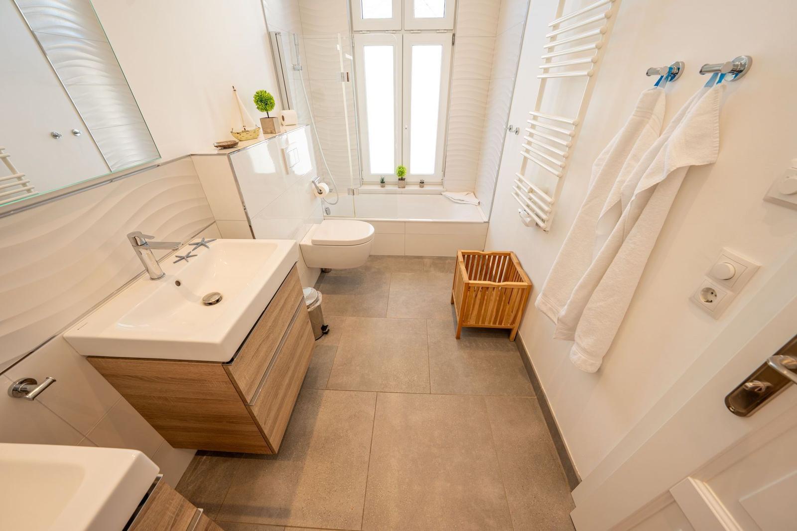 Badezimmer mit Badewanne, Waschbecken und Toilette. Holzregal und Handtücher hängen an der Wand.